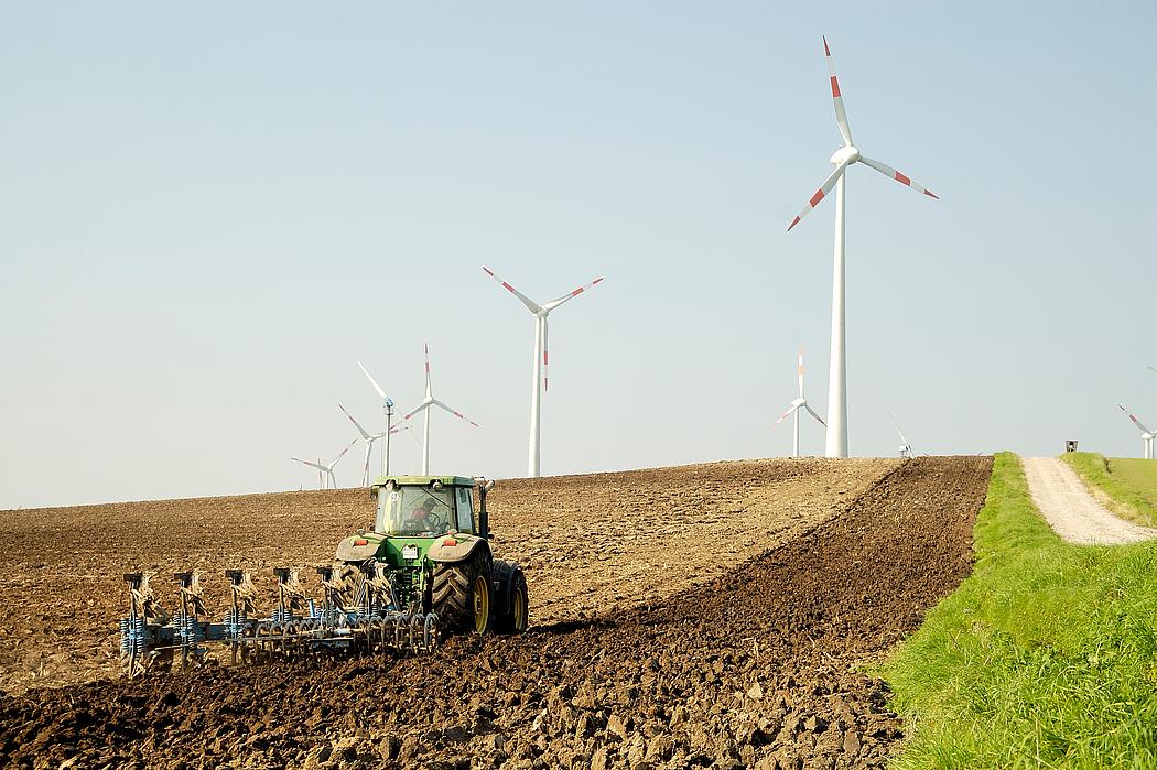 Ein Traktor auf einem Feld. Dahinter ragen Windkraftanlagen in die Höhe.