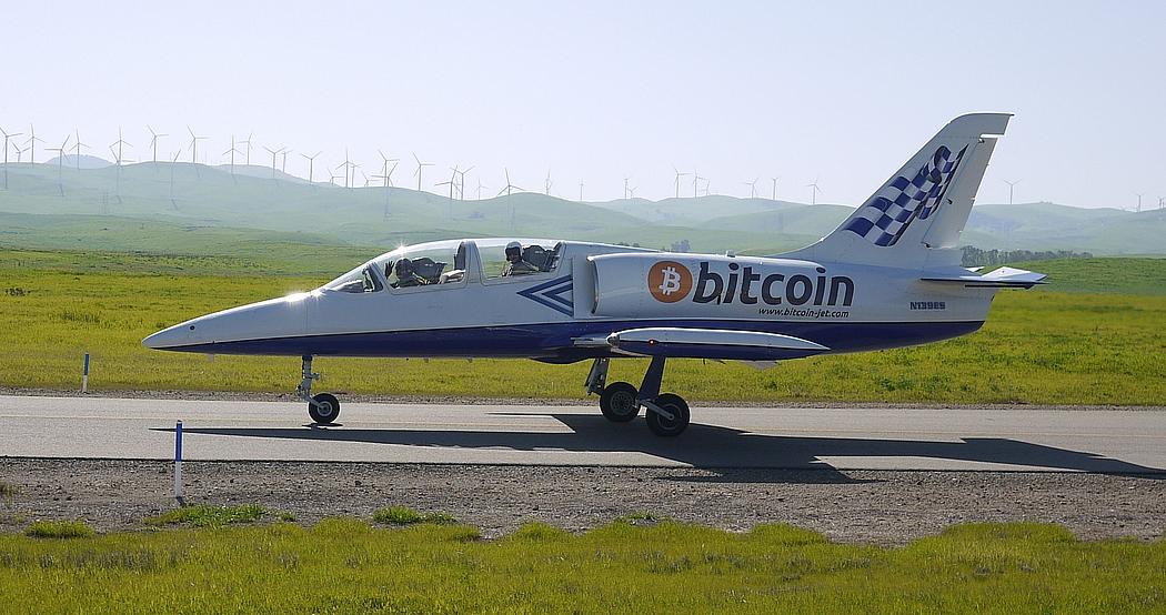 Foto: Bild eines Düsenjets mit der Aufschrift Bitcoin. Im Hintergrund Windräder.