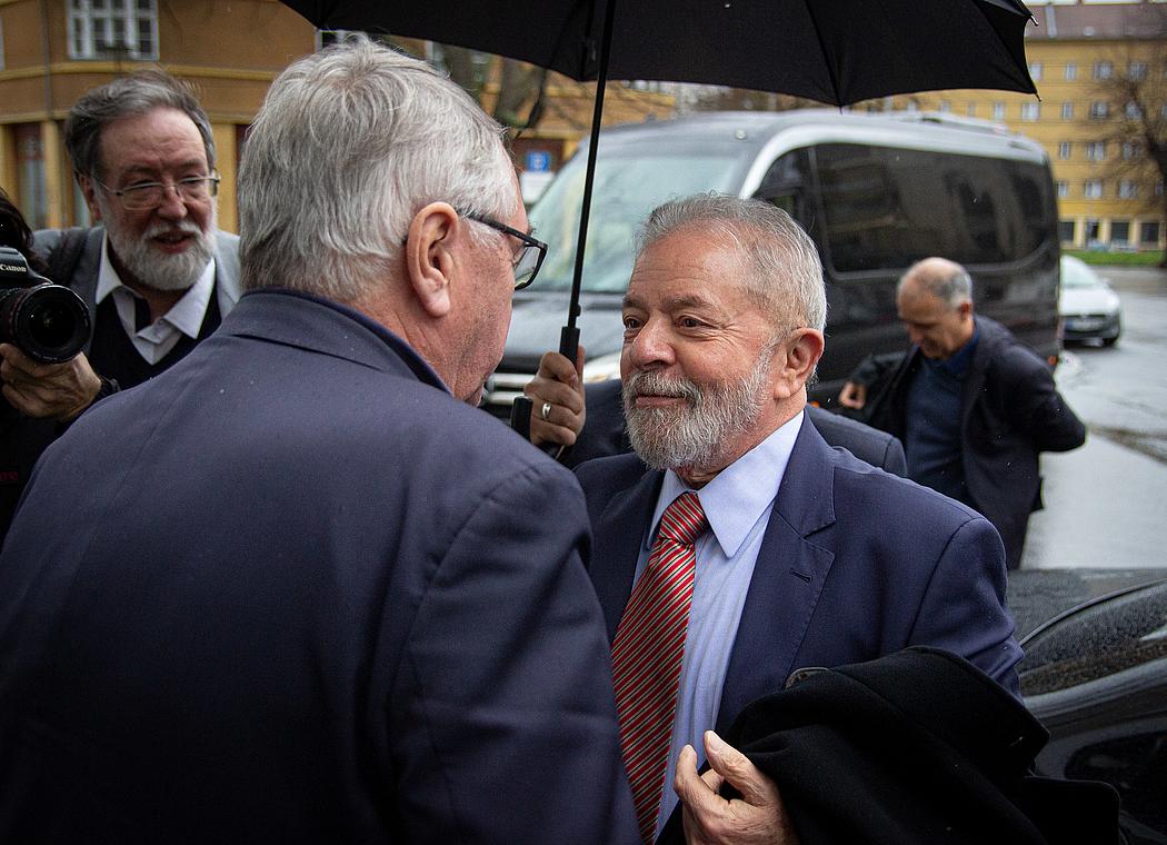 Ein Mann mit grauem Bart und grauen Haaren unter einem Regenschirm, der einen anderen Mann begrüßt