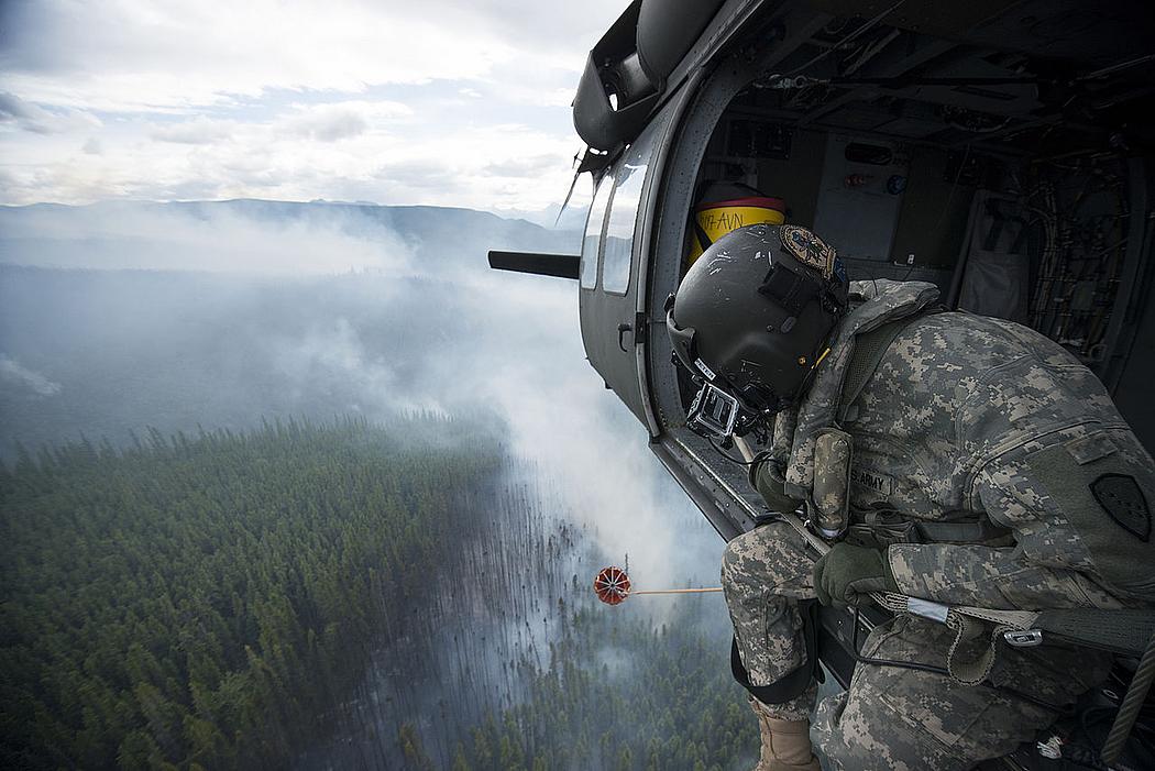 Ein Soldat in Uniform sitzt in einem Helikopter und steuert den Abwurf von Wasser über einem Waldbrand. 