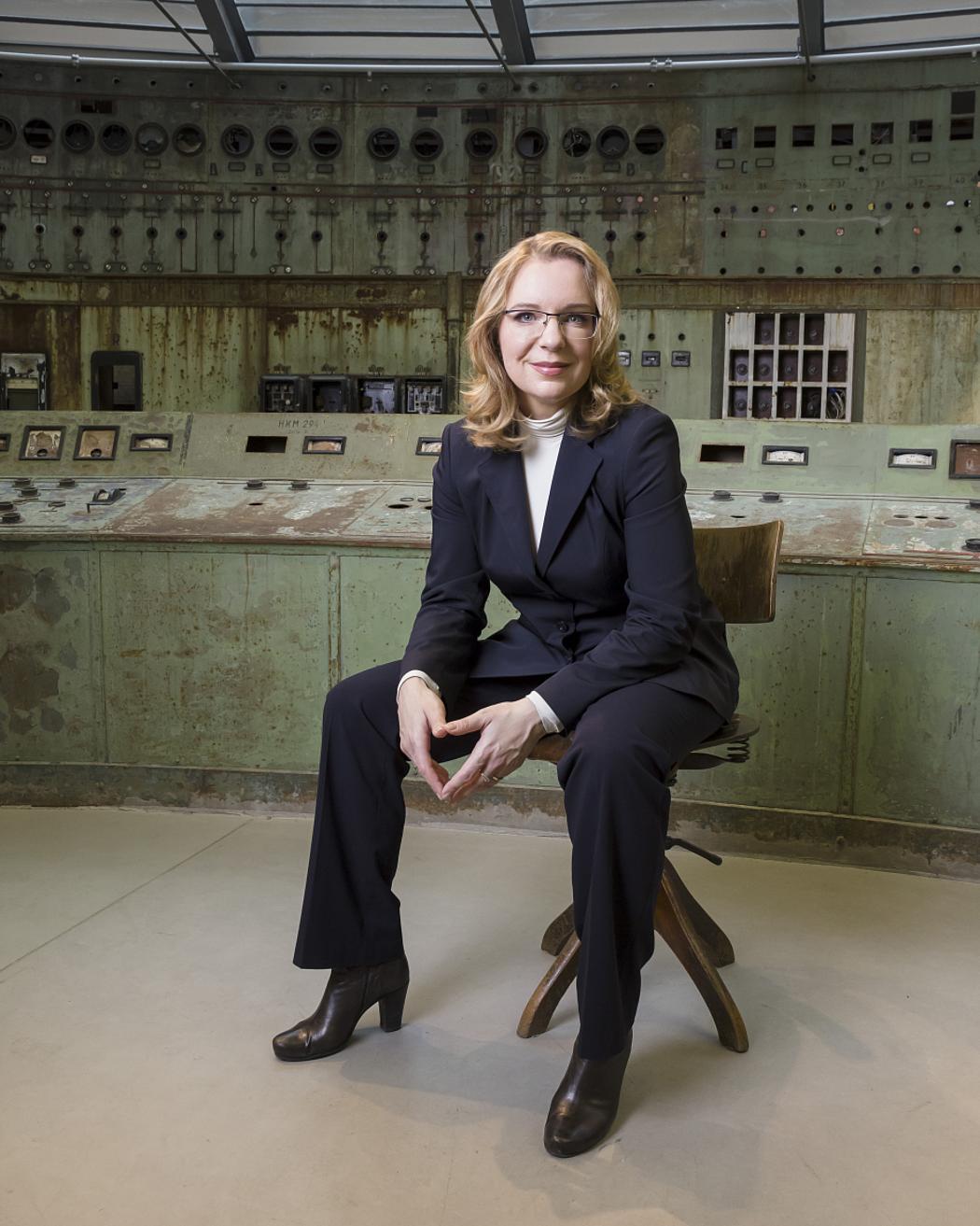 Eine Frau mit blonden Haaren und schwarzem Blazer sitzend in einem alten Kraftwerk