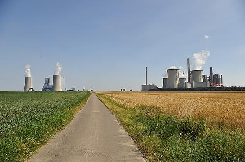 eine kleine Straße und Felder mit mehreren Blöcken eines Kohlekraftwerks links und rechts 