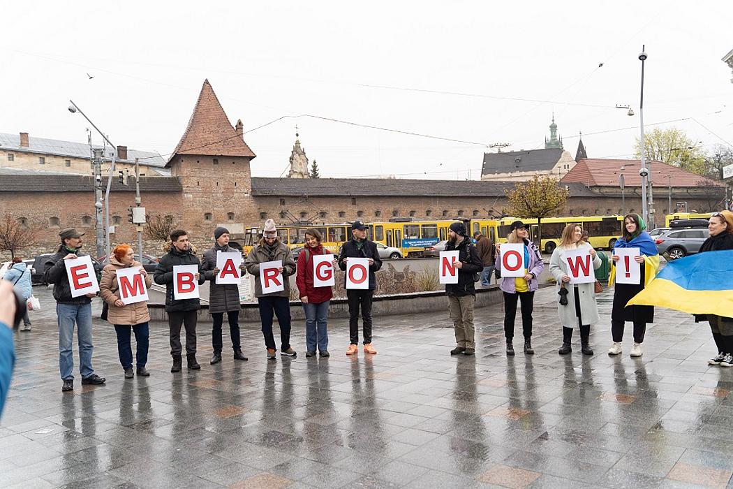 Mehrere Personen halten auf einem Platz Buchstaben hoch, die zusammen "Embargo Now" ergeben. Zudem hält eine Frau eine Flagge der Ukraine. 