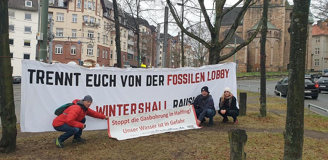 drei Menschen knien vor zwei Plakaten, auf denen steht: 1. "Trennt euch von der fossilen Lobby, Wintershall raus" 2. Stoppt die Gasbohrung in Halfing, unser Wasser ist in Gefahr.