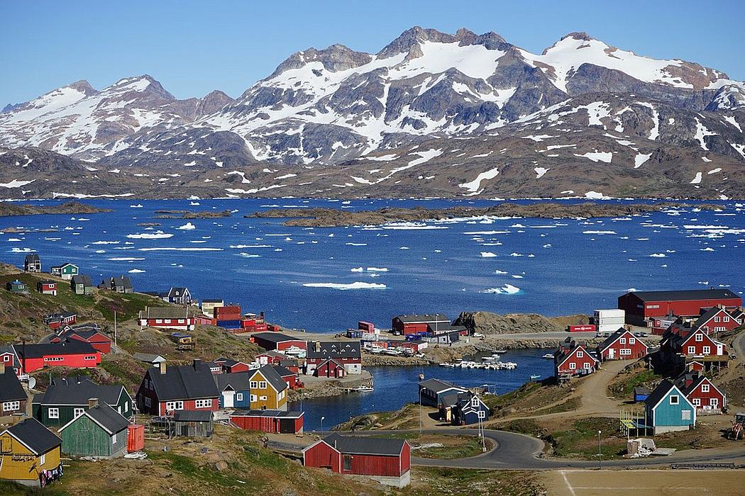 Häuser in Tasiilaq, Grönland, an einem Fjord, im Hintergrund schneebdeckte Berge.