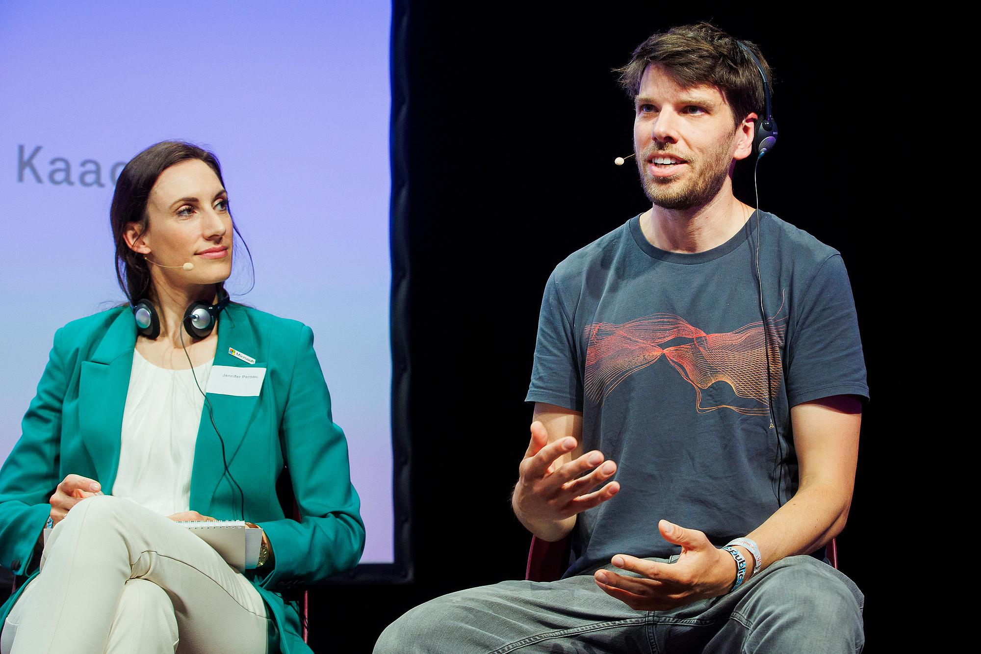 Ein Mann rechts spricht auf einer Bühne. Neben ihm sitzt eine Frau mit weißen T-Shirt und grünem Blazer