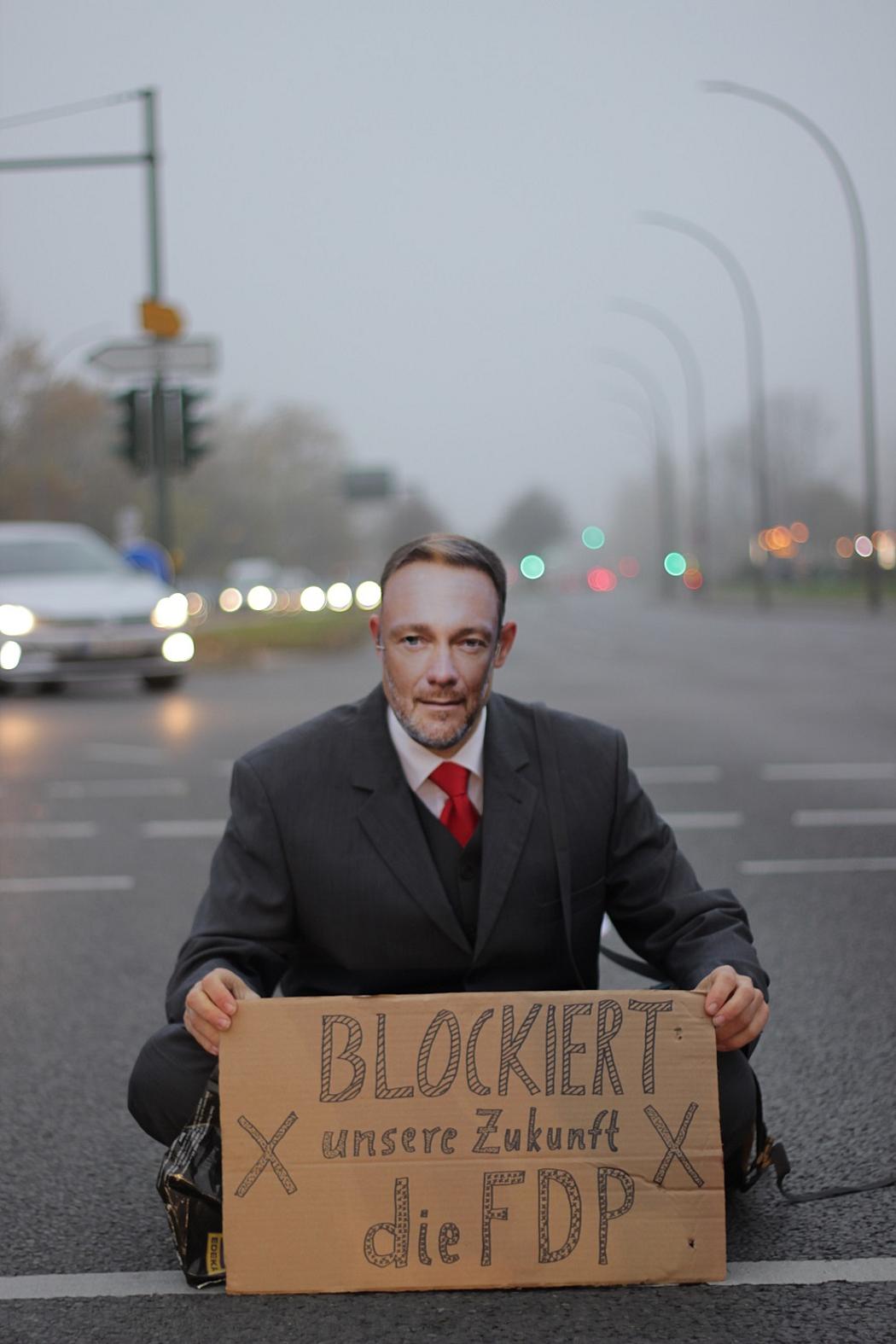 Ein Mann sitzt mit einer Christian-Lindner-Maske und Schild auf einer Straße auf einer 