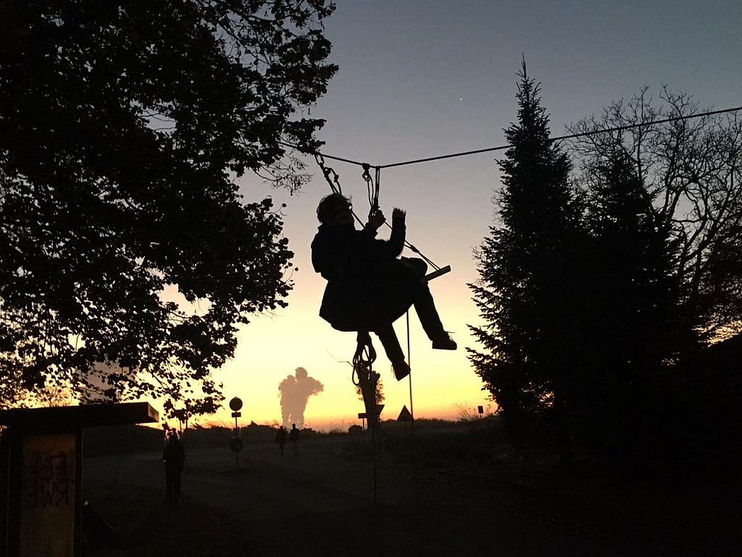 Ein Aktivist hängt an einem Seil über der Straße in der Abenddämmerung von Lützerath. Im Hintergrund sind Rauchsäulen von Kraftwerken zu sehen.