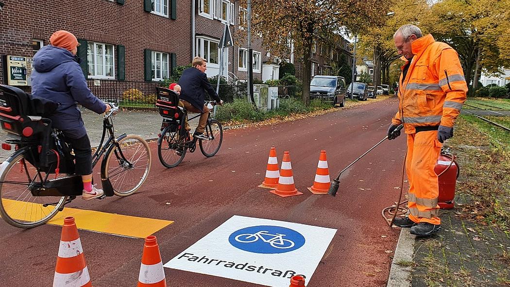 Ein Mann in orangenem Overall bepinselt eine Fahrbahn auf der Fahrradstraße steht. An ihm vorbei fahren Fahrradfahrer.