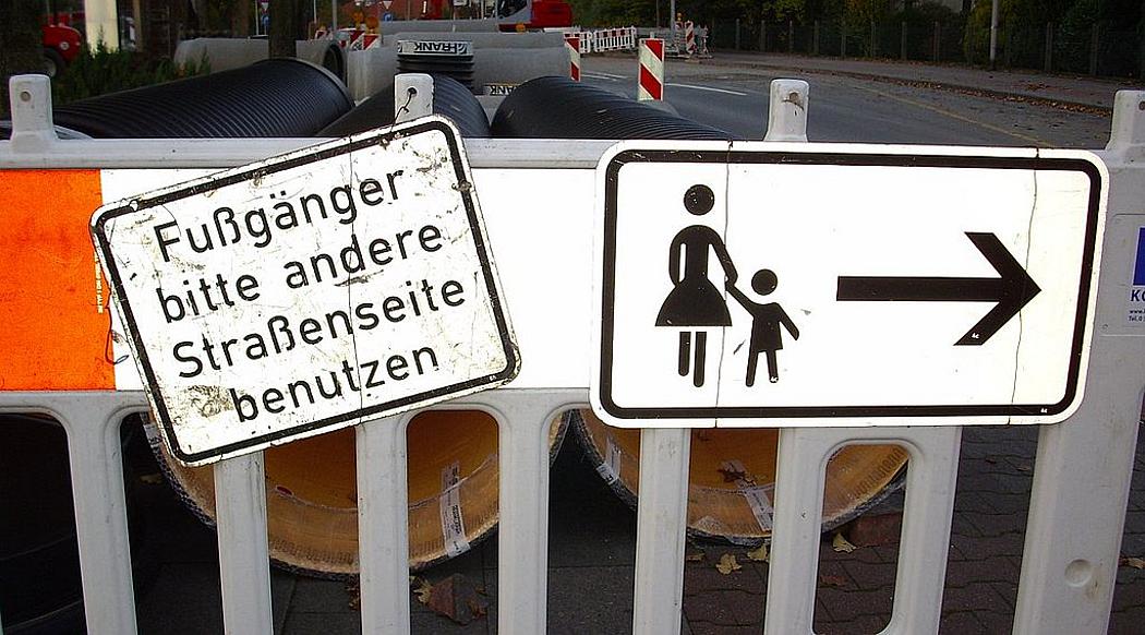Straßenschild vor Baustelle auf dem Gehweg mit der Aufschrift: Fußgänger bitte andere Straßenseite benutzen
