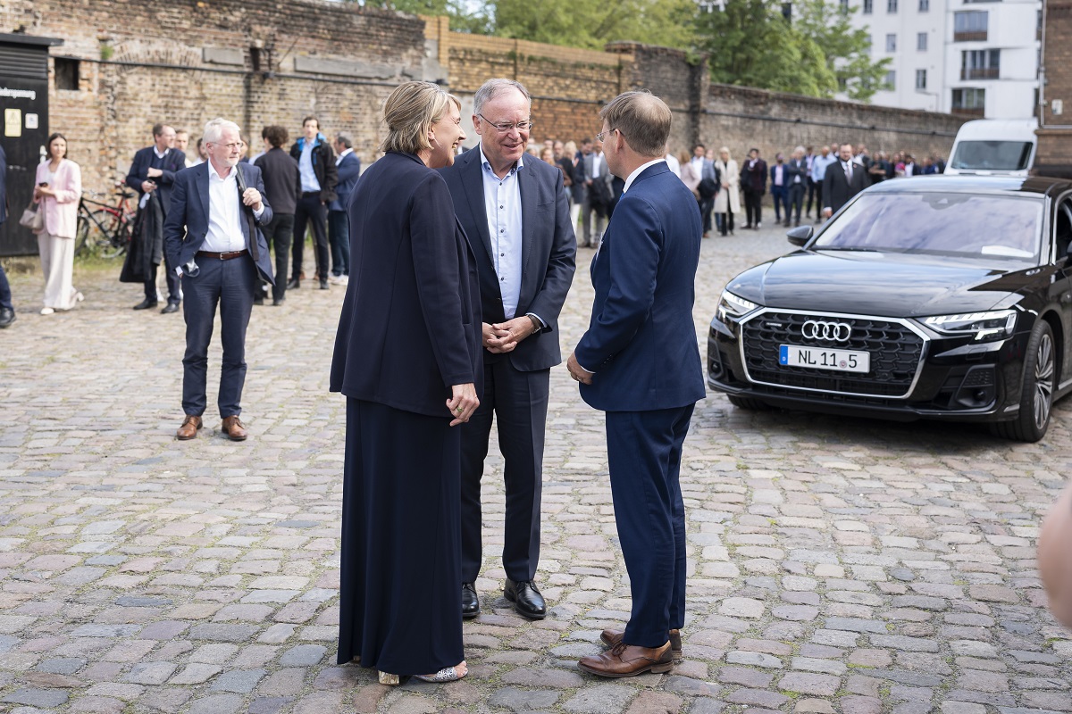 Ein Frau und zwei Männer stehen auf einem Platz. Im Hintergrund ein schwarzes Auto